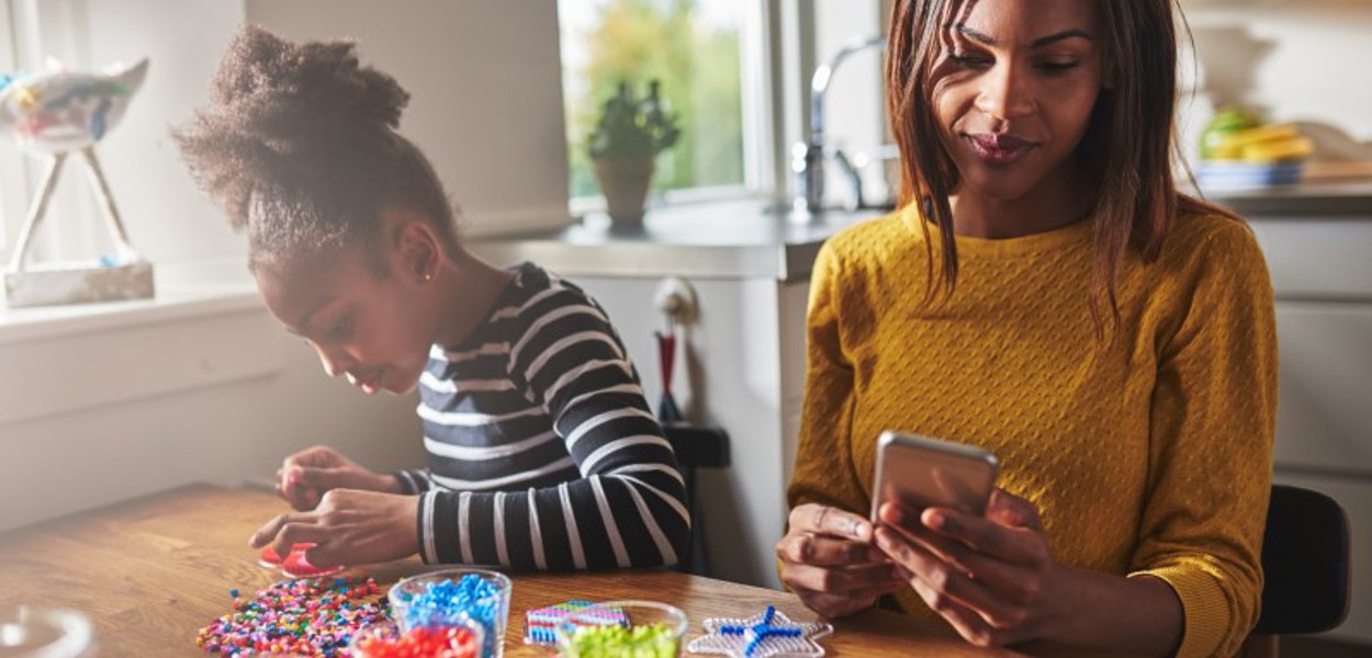 image: a woman looking at her phone sitting at a table while her young daughter does craft activities